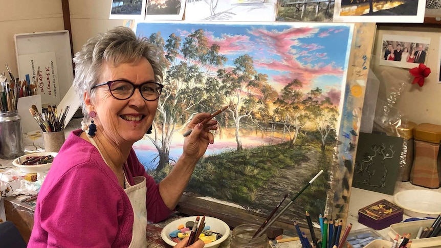 A smiling woman in a pink top and glasses works at a painting on an easel.