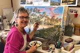 A smiling woman in a pink top and glasses works at a painting on an easel.