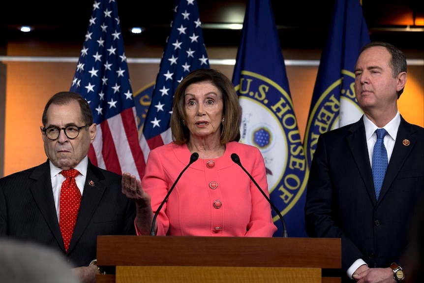 Nancy Pelosi stands at a lectern, with one hand held out in front. Two men in suits stand at her sides.