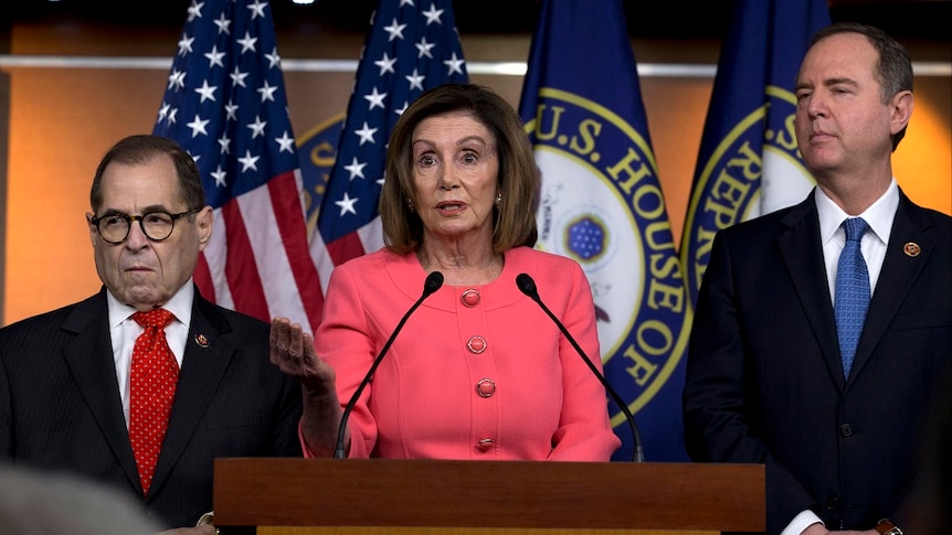 Nancy Pelosi stands at a lectern, with one hand held out in front. Two men in suits stand at her sides.