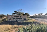 A sign with the words Welcome to Waikerie sits alongside a highway. There is green scrub all around it.