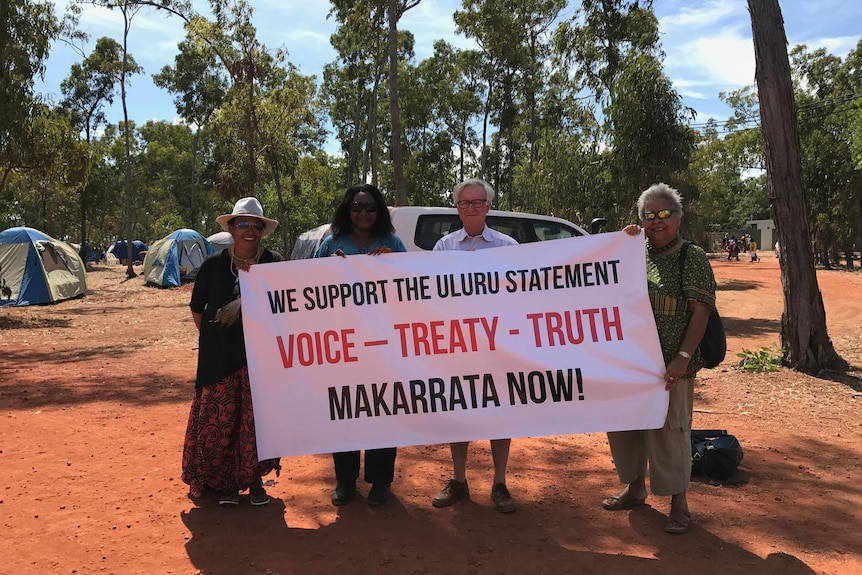 Three Indigenous women and a man with white hair stand in an outback camping ground holding a banner.