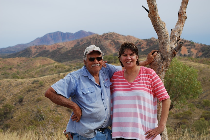 Geoffrey Liddle with his daughter smiling and standing next to a tree, with scrub and mountains in the background.