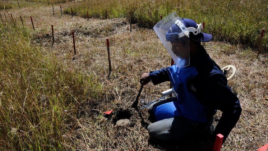 Looking down at a man in blue protective clothing and a full face shield as he works clearing a field of land mines.