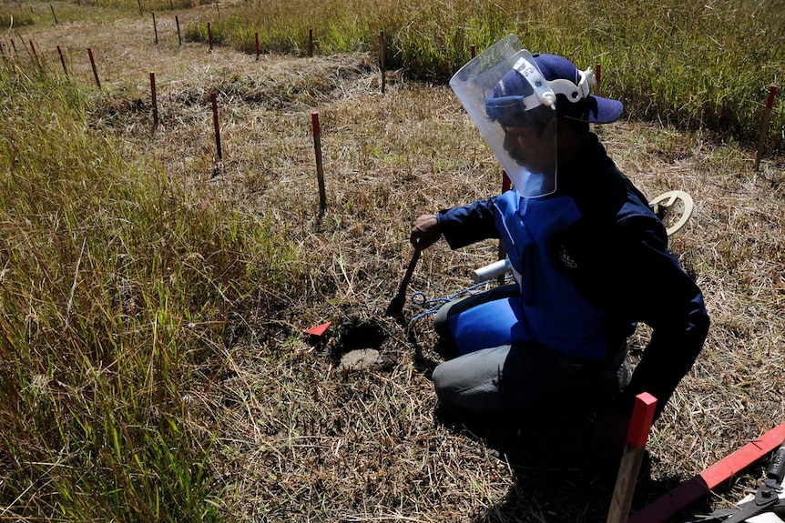 Looking down at a man in blue protective clothing and a full face shield as he works clearing a field of land mines.