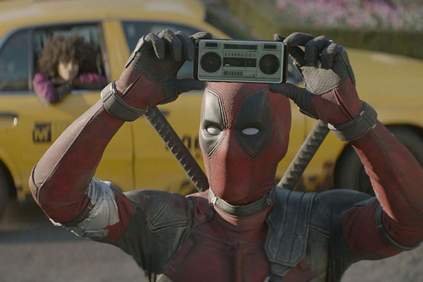 Still image from 2018 film Deadpool 2 of Deadpool standing in front of yellow taxi, holding up an image of a boombox on a phone