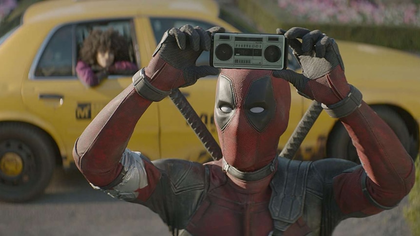 Still image from 2018 film Deadpool 2 of Deadpool standing in front of yellow taxi, holding up an image of a boombox on a phone