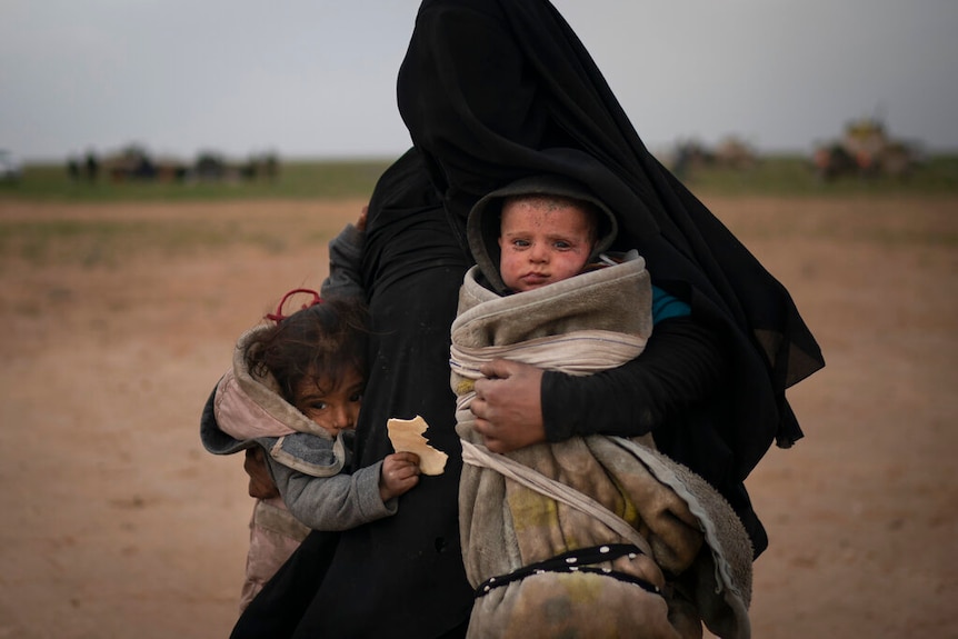 A woman wearing a black niqab holds a baby swaddled in a blanket, while a toddler clings to her side. The trio are in a desert.