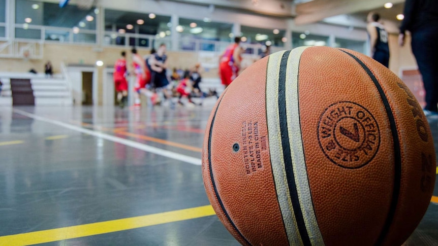 An orange and white basketball lies on the court in a gym with a game being played in the background.