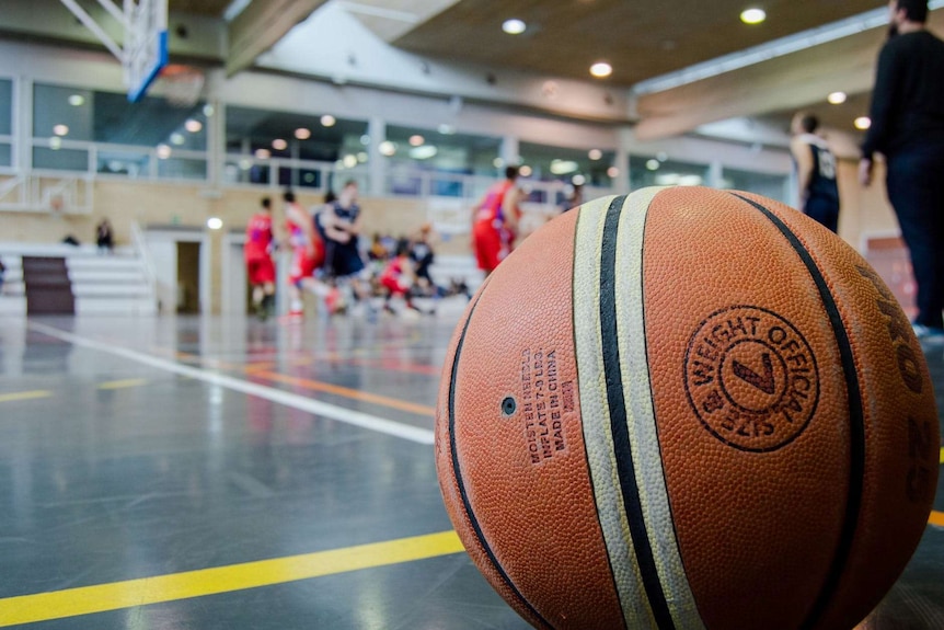 An orange and white basketball lies on the court in a gym with a game being played in the background.