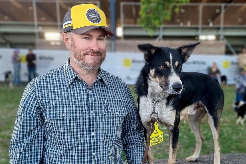 A man in a yellow cap smiling and holding the lead of a dog next to him