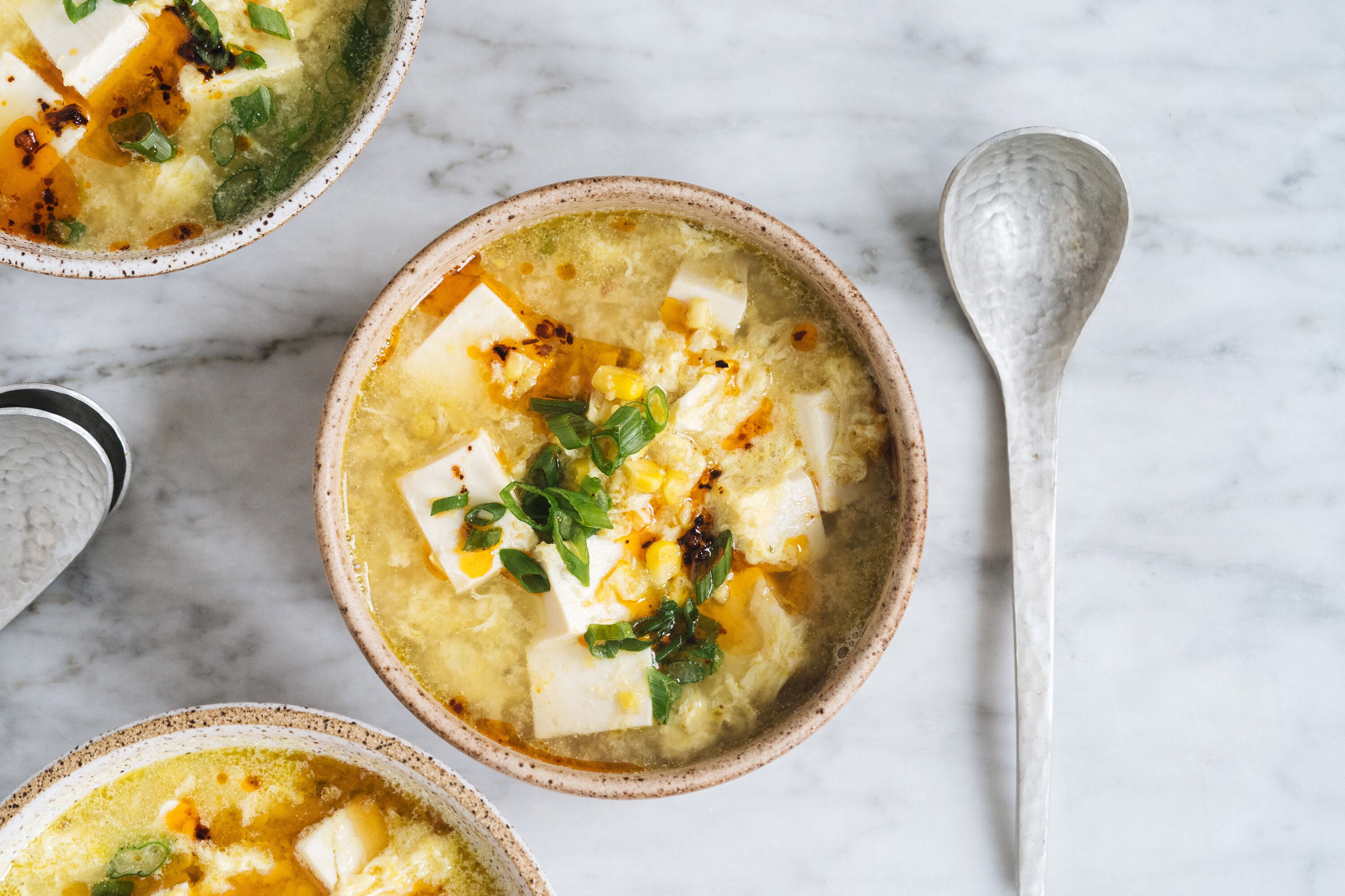A bowl of tofu and sweetcorn soup with strands of egg, topped with spring onion and chilli oil, a warming vegetarian dinner.