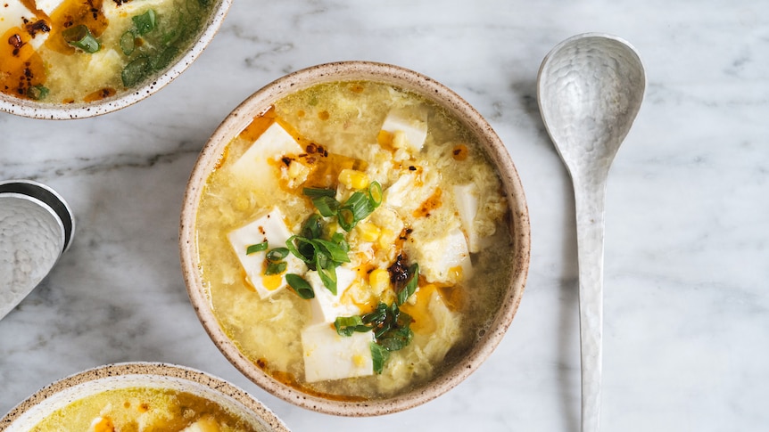A bowl of tofu and sweetcorn soup with strands of egg, topped with spring onion and chilli oil, a warming vegetarian dinner.