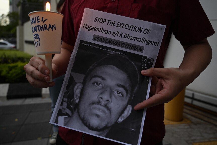Man holds a cup with a candle inside it and a poster that reads 'stop the execution of Nagaenthran K. Dharmalingam' 