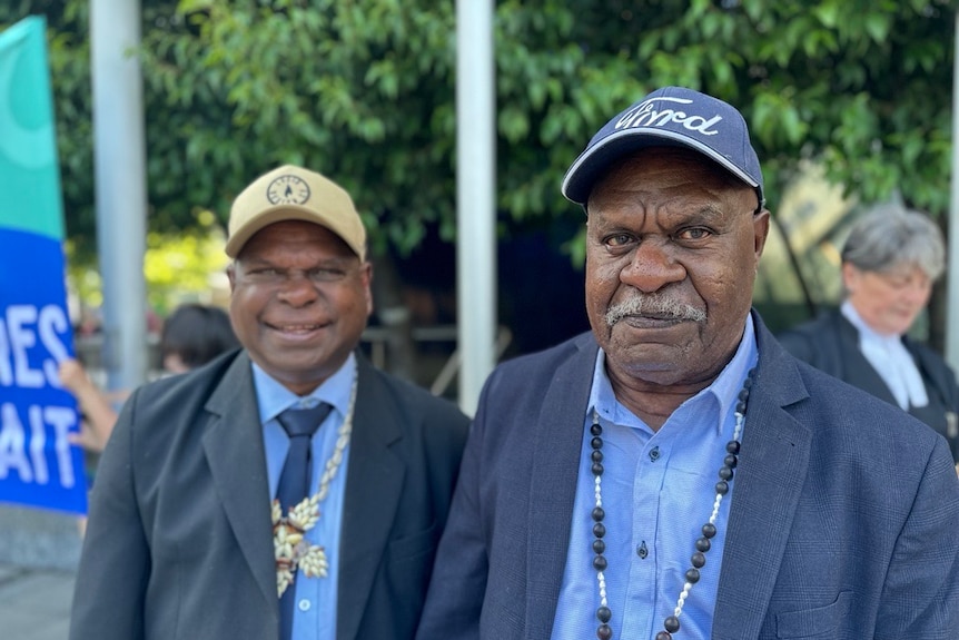 Two indigenous men in suits and caps smile into the camera. 