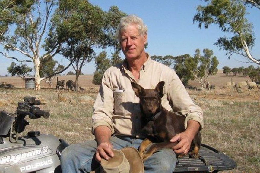 White, middle-aged man with grey hair, wearing a shirt and sitting on a quad bike and a red kelpie dog