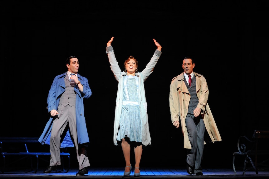 A woman on stage with her hands in the air, there is a man either side of her, mid-dance move