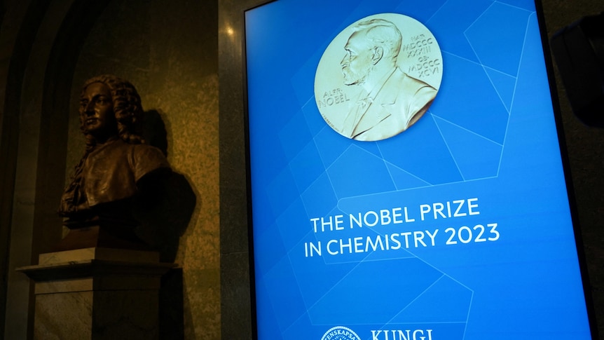 A bronze bust stands next to a TV screen displaying a title card for the 2023 Nobel Prize in Chemistry.