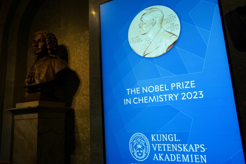A bronze bust stands next to a TV screen displaying a title card for the 2023 Nobel Prize in Chemistry.