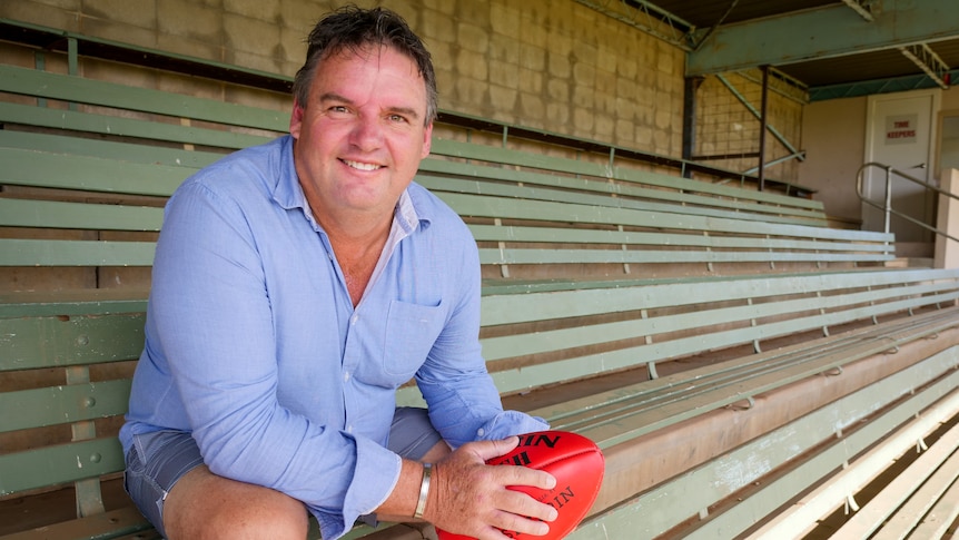 A man wearing shorts and a blue shirt sitting in a grandstand holding an AFL football.