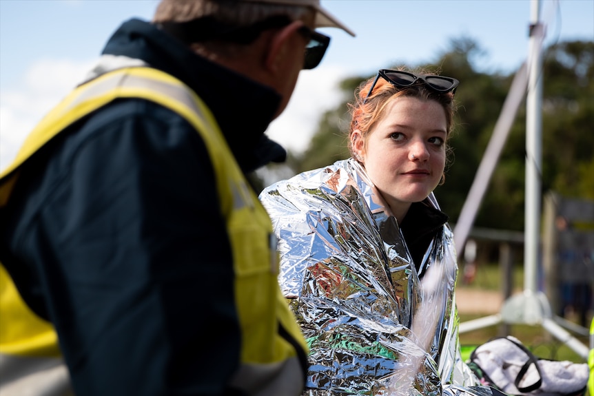 A woman in an emergency blanket with cup of tea,sunnies on top, looks at a man in a high viz jacket.