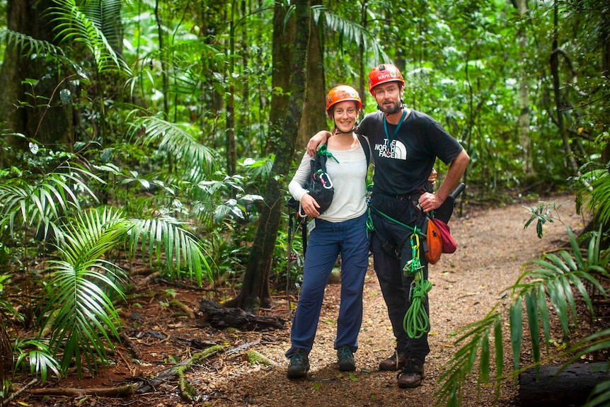 A man and woman with arms around in each other, wearing helmets and carrying climbing equipment, standing on a path in a forest.