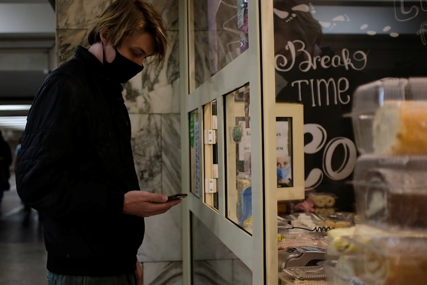 A man stands outside a bakery window where pastries and cakes are on display. He is looking at his phone