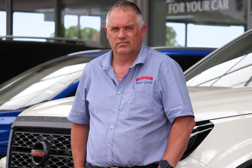 a man with short, grey hair, blue short sleeved shirt stands in front of an office with parked cars