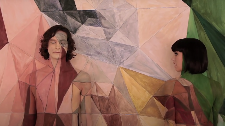Gotye and Kimbra are topless and painted the same colours and pattern as the wall behind them