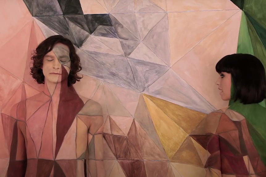 Gotye and Kimbra are topless and painted the same colours and pattern as the wall behind them