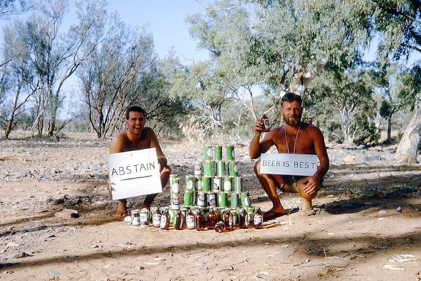 Two men holding signs saying 'abstain' and 'beer is best' sitting next to a pile of beer bottles and cans.