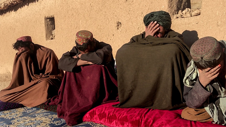 Three men and a young boy, sitting in robes and visibly grief stricken, sitting in front of a compound.