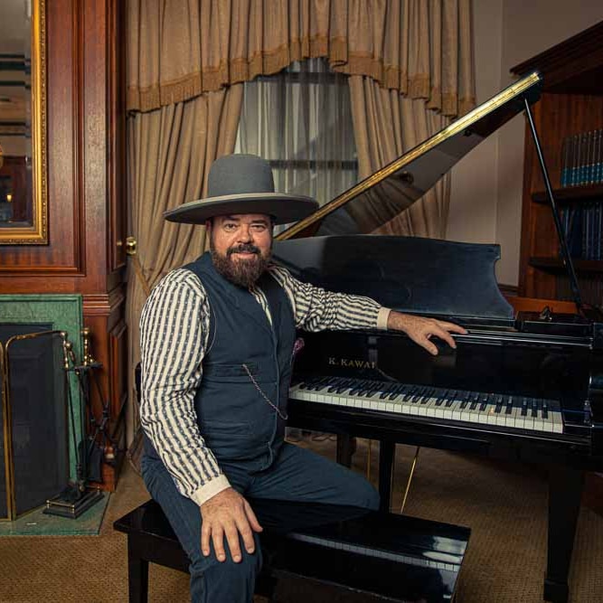 Andrew Farriss of INXS sits at a grand piano in a dining room, wearing country attire.