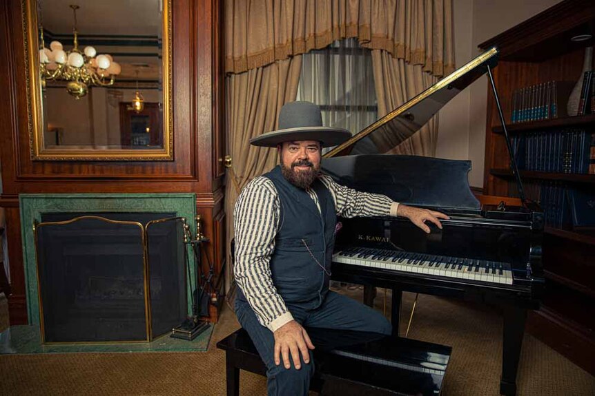 Andrew Farriss of INXS sits at a grand piano in a dining room, wearing country attire.
