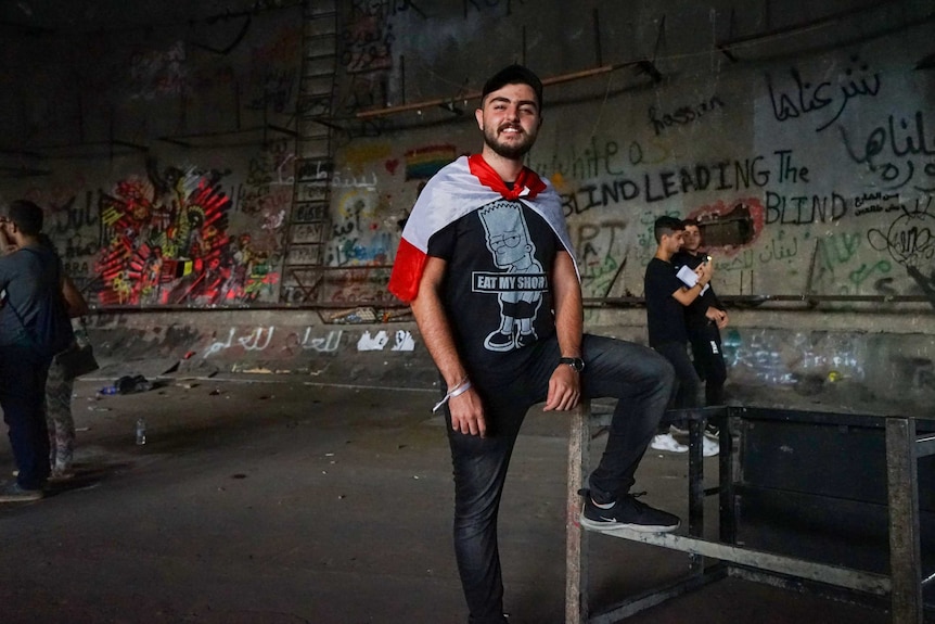 A young man in a Simpsons t-shirt wrapped in a Lebanese flag stands in front of a wall covered in graffiti