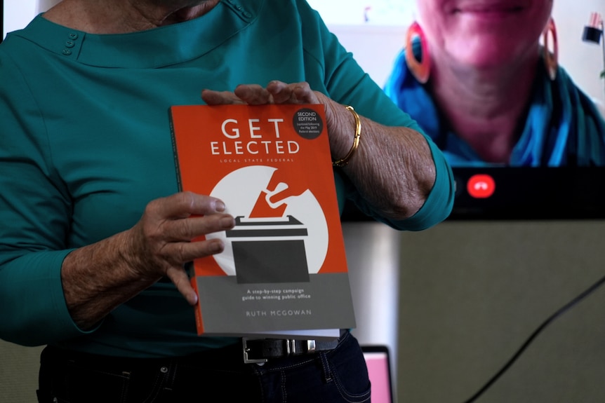 A woman wearing a teal shirt holds a book with orange cover that reads Get Elected. Her face is not visible.