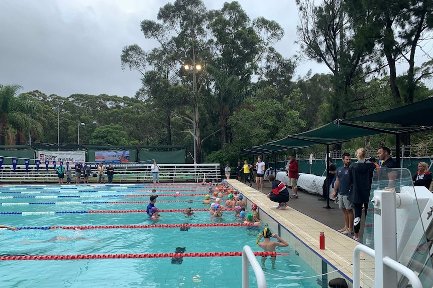 Kids in a swimming pool as adults look on and one person coaches from the side.