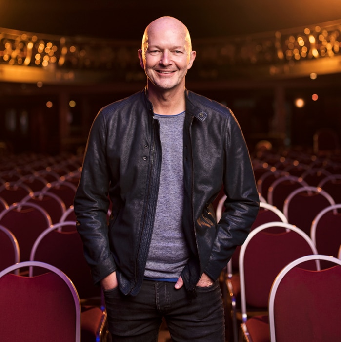 David Berthold inside a theatre, standing amongst red seats and backlit with orange spotlights.