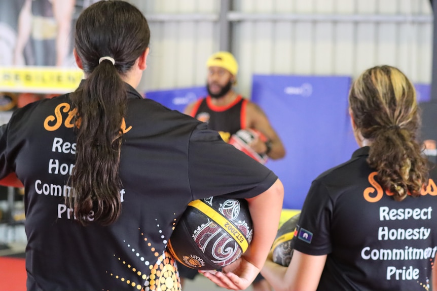 Two young Indigenous girls, back to the camera, holding basketballs and watching someone talk to them inside a sports gymnasium.