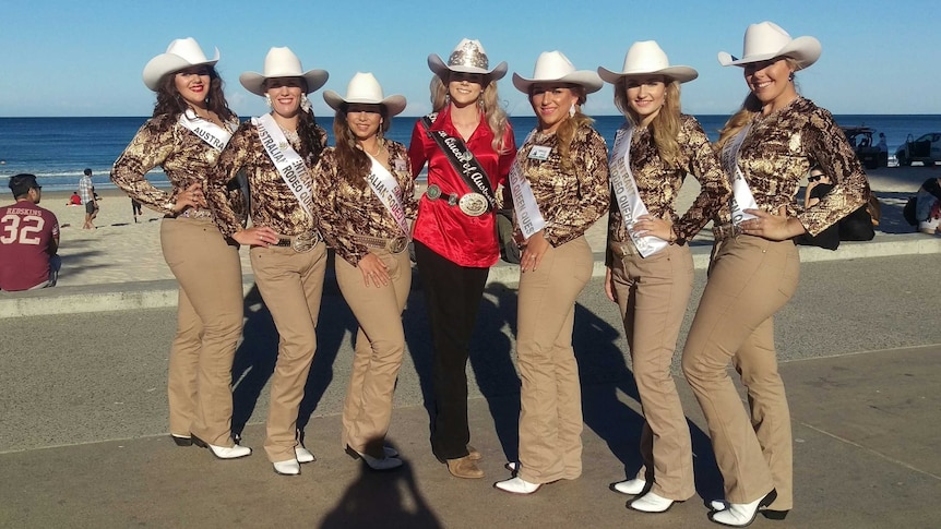 Seven girls wearing cowboy hats, brown pants and patterned shirts stand near a beach.