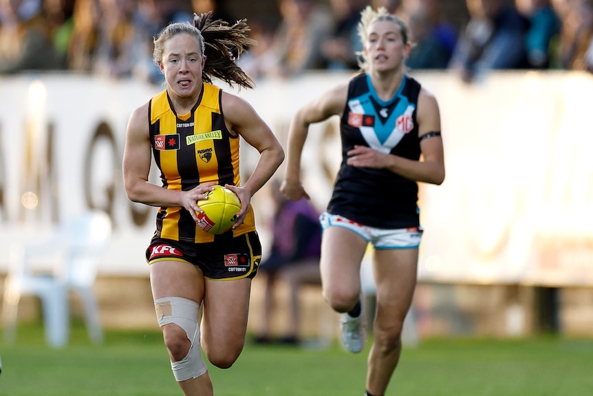 A Hawthorn AFLW player runs with the ball as a Port Adelaide opponent gives chase.
