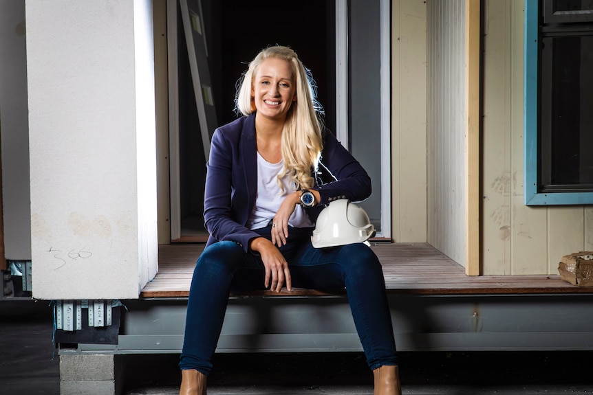 Blonde woman smiling and sitting on the deck of a small home with a hard hat resting on her knee