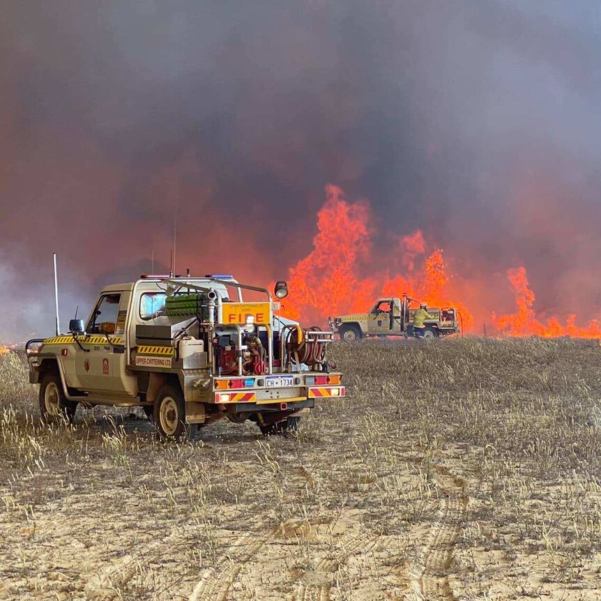 Two fire trucks pictured on an intense fire front with red flames right in front of one of the trucks