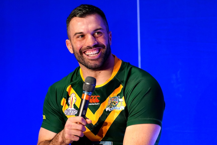 Dressed in his Australian Kangaroos jersey, James Tedesco laughs while holding a microphone in front of a blue background.