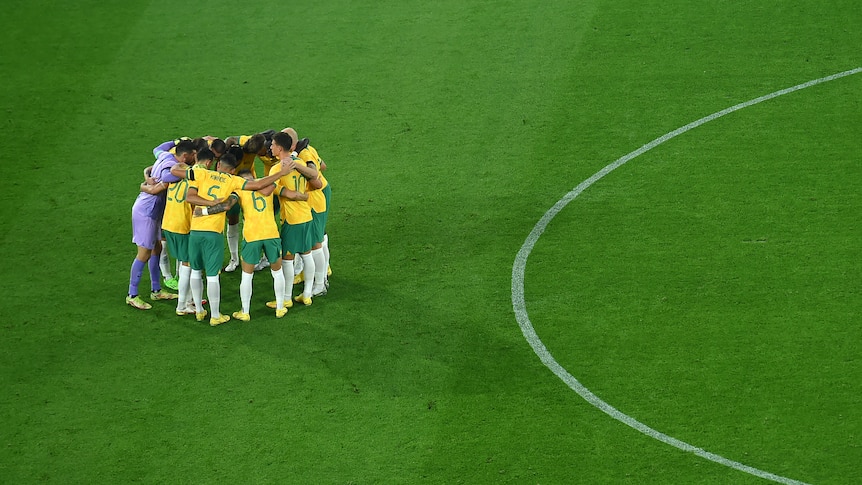 A male soccer team wearing yellow and green stands in a circle on some green grass