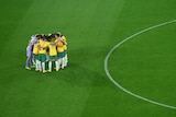 A male soccer team wearing yellow and green stands in a circle on some green grass