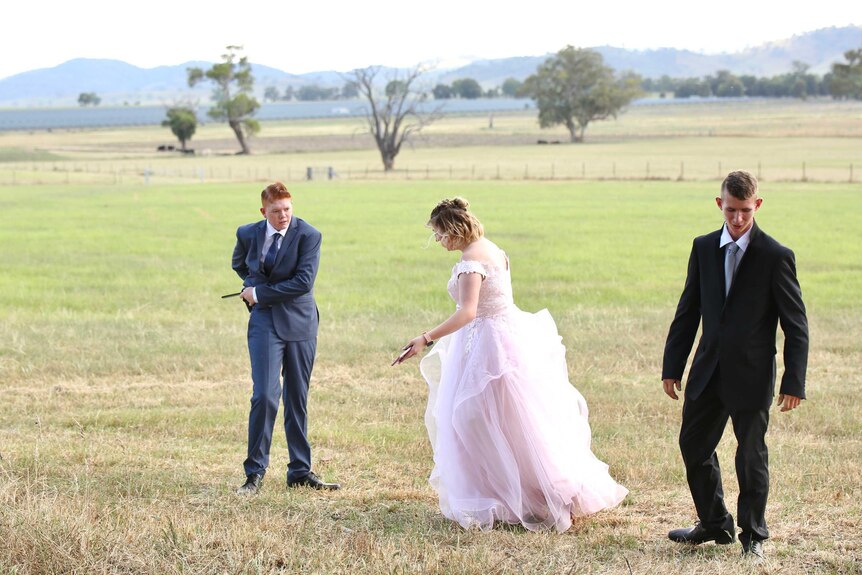 Three year 12's in formalwear stand in a paddock. 1 men in suits. 1 woman in a big puffy pink dress.