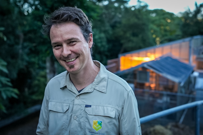 A man stand in front of a well-lit wooden house at dawn, smiling at the camera. 