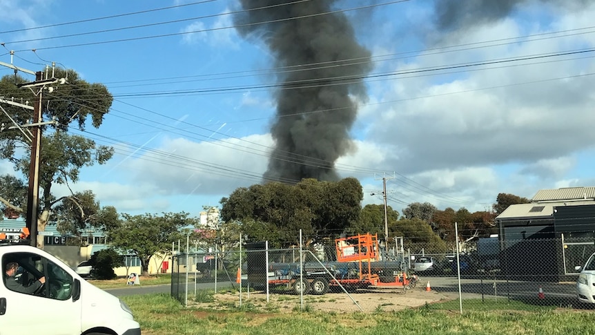 Black smoke rising in a large plume above an industrial area, taken from a street several blocks away