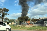 Black smoke rising in a large plume above an industrial area, taken from a street several blocks away